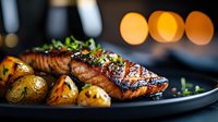 Grilled salmon fillet with roasted potatoes and fresh herbs on dark plate in restaurant setting with warm bokeh lights background.