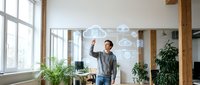 Young businessman interacting with floating cloud computing icons in modern office space with wooden beams and large windows for technology presentations.