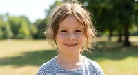 Happy young Caucasian girl with blonde hair smiling outdoors in sunny park setting wearing striped shirt for childhood and family lifestyle concepts.