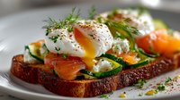 Gourmet breakfast toast with poached egg, smoked salmon, zucchini slices, and fresh dill garnish on rustic bread, served on white plate.