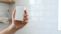 Male hand holding blank white rectangular box or package against modern kitchen background with white subway tile wall, suitable for product mockup, packaging design, and commercial advertising.