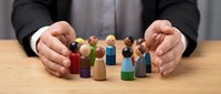 Businessman in dark suit protecting diverse wooden peg dolls in various colors on table, representing team unity, diversity, and employee care in corporate environment.