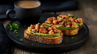Fresh salmon and avocado toast on dark seeded bread garnished with herbs and chives, served with coffee on black plate against rustic wooden background.