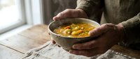 Hands holding ceramic bowl with warm corn soup or chowder, rustic wooden table setting, comfort food meal preparation.