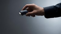 Close-up of male hand in black sleeve holding small black USB flash drive against neutral gray background for technology and data storage concepts.