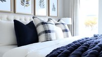 Cozy modern bedroom interior with navy blue and plaid pillows, chunky knit blanket, and botanical wall art. Natural light from window creates serene atmosphere.