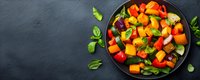 Fresh colorful roasted vegetables with pumpkin, bell peppers, zucchini and basil leaves served on black plate against dark background, top view.
