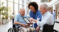 African American female healthcare worker in blue scrubs showing tablet to two elderly Caucasian men in modern medical facility lobby with natural lighting.