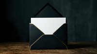 Elegant black envelope with gold trim and blank white card on wooden surface, dark moody background, luxury stationery concept.