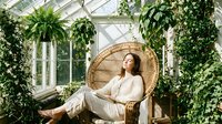 Young Asian woman relaxing in wicker peacock chair surrounded by lush green plants in bright sunlit greenhouse conservatory for wellness and mindfulness.