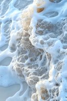 Foamy ocean waves washing over sandy beach shore with white sea foam bubbles and water texture for coastal summer vacation backgrounds.