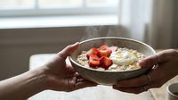 Hands holding bowl of oatmeal porridge topped with fresh strawberries and almonds near window, healthy breakfast meal for nutrition and wellness content.