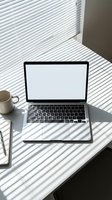 Modern laptop with blank white screen on desk with coffee cup and notebook, natural sunlight creating shadow patterns through window blinds for remote work setup.