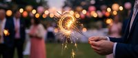 Cropped hand holding lit sparkler at outdoor evening celebration with blurred crowd and bokeh lights in background, suitable for festive event marketing and holiday promotions.