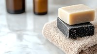 Natural handmade soap bars in black charcoal and beige colors stacked on soft towel with essential oil bottles in background, spa wellness concept.