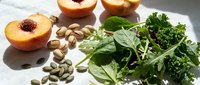 Fresh peach halves with pumpkin seeds and mixed green leafy vegetables including spinach and kale arranged on white surface for healthy eating concept.
