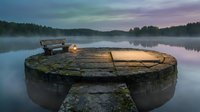 Serene lake at dawn with wooden dock and lantern creating peaceful morning atmosphere. Misty water surface reflects colorful sky in tranquil natural setting.
