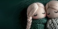 Two handmade textile dolls with closed eyes and braided hair snuggling together, wrapped in cozy knitted green and gray sweaters against dark background.