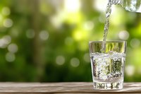 Fresh pure water pouring into clear glass on wooden surface against blurred green natural background with bokeh lights, refreshing drink concept.