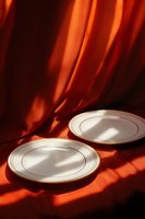 White porcelain plates with gold trim on burnt orange silk fabric backdrop with dramatic shadows and light creating elegant dining atmosphere.