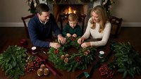 Caucasian family making Christmas wreath together at home with fireplace, holiday crafting activity with parents and child decorating festive garland.