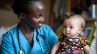 African American female healthcare provider in blue scrubs with stethoscope holding smiling Caucasian baby in colorful floral outfit during pediatric checkup.