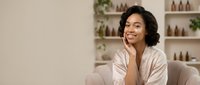 Young woman with dark wavy hair and medium skin tone, wearing a light pink button-up shirt, sitting on a beige chair in a modern spa or wellness clinic interior with shelves of beauty products and plants behind her, smiling thoughtfully while touching her face.