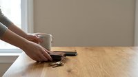 Woman hands holding white coffee mug on wooden table near window in minimalist home interior for morning routine and relaxation concepts.