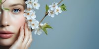 Young woman with natural makeup and blue eyes holding cherry blossom branch against blue background, representing spring beauty and skincare concept.