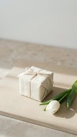 Elegant gift box wrapped in natural linen fabric with cream ribbon bow alongside fresh white tulip on neutral beige surface for special occasions.