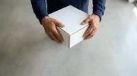 Man holding white cardboard box with both hands, wearing dark blue shirt, overhead view on concrete surface for shipping, delivery, and packaging concepts.
