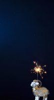 Decorative white and gold ram figurine holding a sparkling sparkler against dark blue night sky background, suitable for holiday celebrations, festive greeting cards, and seasonal marketing materials.