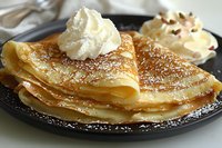 Delicate crepes folded on black plate dusted with powdered sugar, topped with fresh whipped cream and garnished with chocolate shavings, served for breakfast.