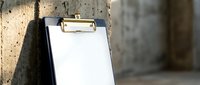 Empty clipboard with blank white paper leaning against concrete wall in industrial setting for business planning and note-taking applications.