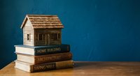 Miniature wooden house model sitting on stack of architecture and home building books against blue background for real estate education concept.