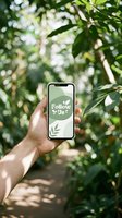 Hand holding smartphone with Follow Us green leaf social media screen in lush tropical garden path, nature eco brand promotion concept.