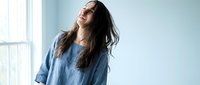 Young woman with long dark hair laughing joyfully in blue casual shirt by bright window, expressing happiness and positive emotions in modern interior.