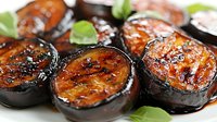 Grilled eggplant slices glazed with sweet soy sauce and garnished with fresh basil leaves, close-up view showing caramelized surface and appetizing char marks.