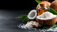 Fresh coconut flakes in halved shells with organic oil and green tropical leaves on dark moody background, spa wellness and natural cosmetics concept.