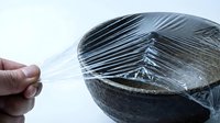 Closeup of hand stretching transparent plastic wrap over wooden bowl against white background, food storage and kitchen preparation concept.