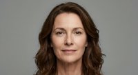 Professional middle-aged Caucasian businesswoman with wavy brown hair smiling confidently against neutral gray background for corporate headshots.