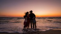 Family of four silhouetted against golden sunset on ocean beach, parents and children embracing during romantic evening vacation moment by waves.