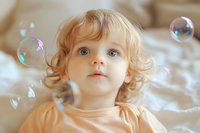 Caucasian toddler with blonde curly hair and blue eyes gazes up in wonder at floating soap bubbles. Soft natural lighting creates dreamy atmosphere for childhood innocence.