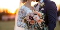 Romantic sunset wedding moment with bride holding blush pink rose bouquet, lace dress detail, and groom in navy suit embracing during golden hour photography session.
