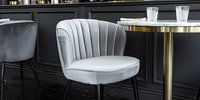 Elegant light gray velvet shell chair with vertical channel tufting and brass base against dark wall in modern restaurant setting. Round bistro table with metallic accents.