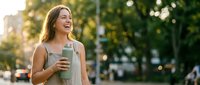 Young Caucasian woman laughing outdoors, holding reusable water bottle in sunny urban park, embodying eco-friendly lifestyle and wellness.
