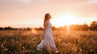 Woman in white lace dress standing in blooming wildflower field at golden hour sunset. Serene outdoor portrait for lifestyle, fashion, and nature themes.