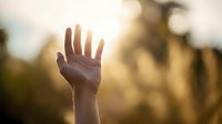 Hand reaching toward golden sunset light with blurred natural background, creating spiritual atmosphere for concepts of hope, faith, and connection.