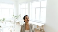 Woman with dark hair and medium skin tone wearing a beige cardigan in a bright, minimalist home office with large windows, potted plants, and wooden furniture, looking upward thoughtfully.
