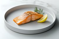 Grilled salmon fillet seasoned with herbs and spices on round gray ceramic plate with fresh dill and lemon slice, served on white marble surface.
